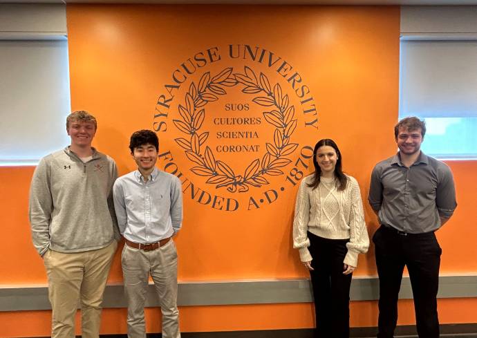 four people standing against a wall with the Syracuse University seal