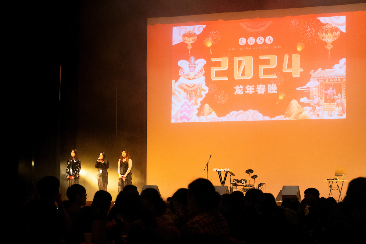 Three people on stage with a large screen in the background and Chinese writing on it. 