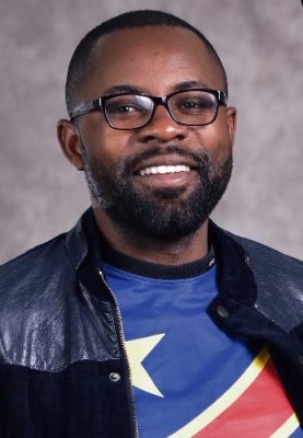 Person wearing a leather-sleeved jacket over a shirt featuring a bold, colorful star and diagonal stripe design, posed against a neutral studio backdrop.