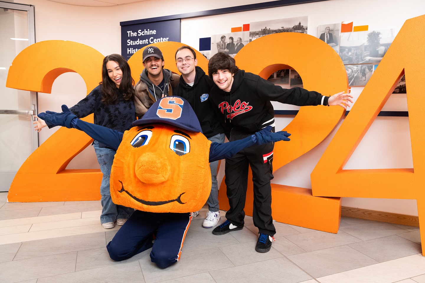 four people standing behind Otto the Orange in front of large set of numbers of 2024