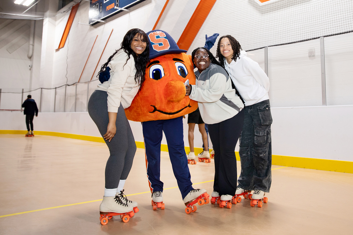 Three students on roller skates pose for a photo with Otto the Orange.