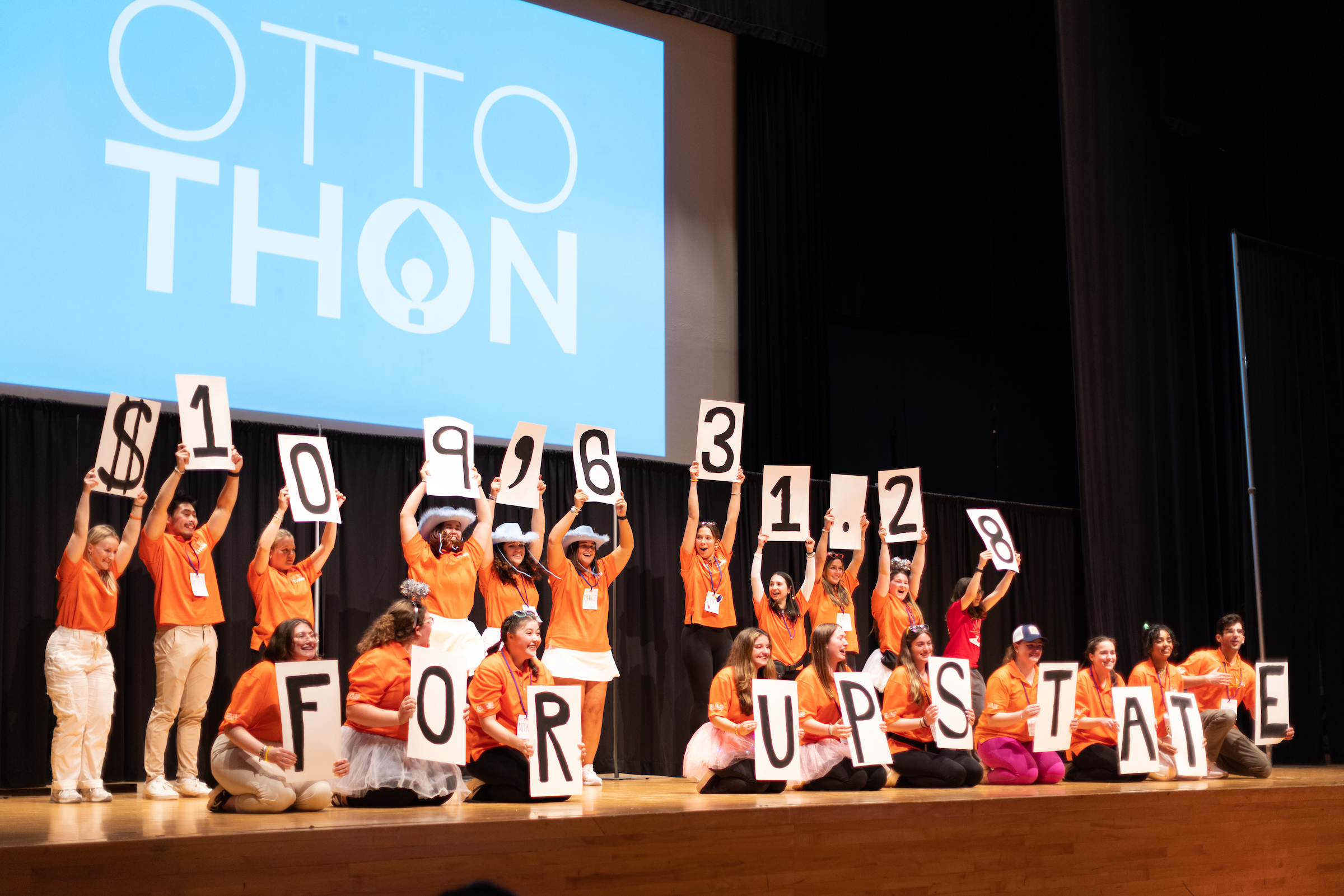 group of people sitting and standing on stage holding up placards that represent $109,631.28 and For Upstate, in front of projection screen with text OttoThon