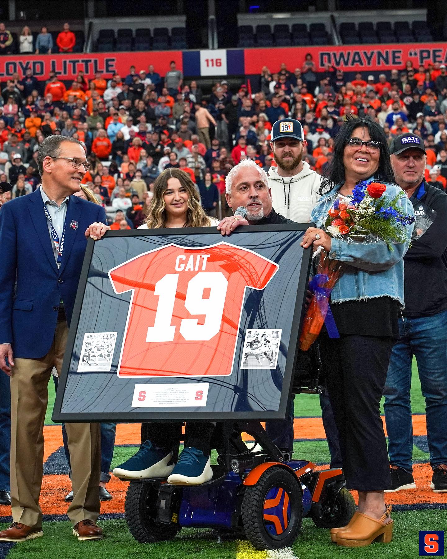 Man in wheelchair holding a framed orange 19 jersey with Gait written above the number surrounded by people.