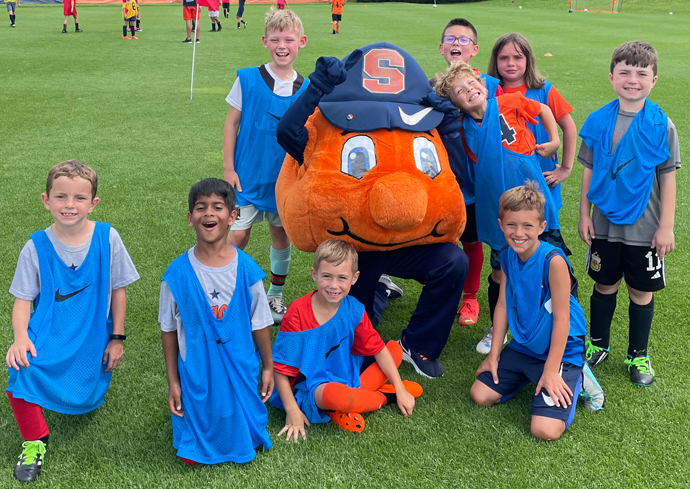 Children pose with Otto the Orange on a soccer field.