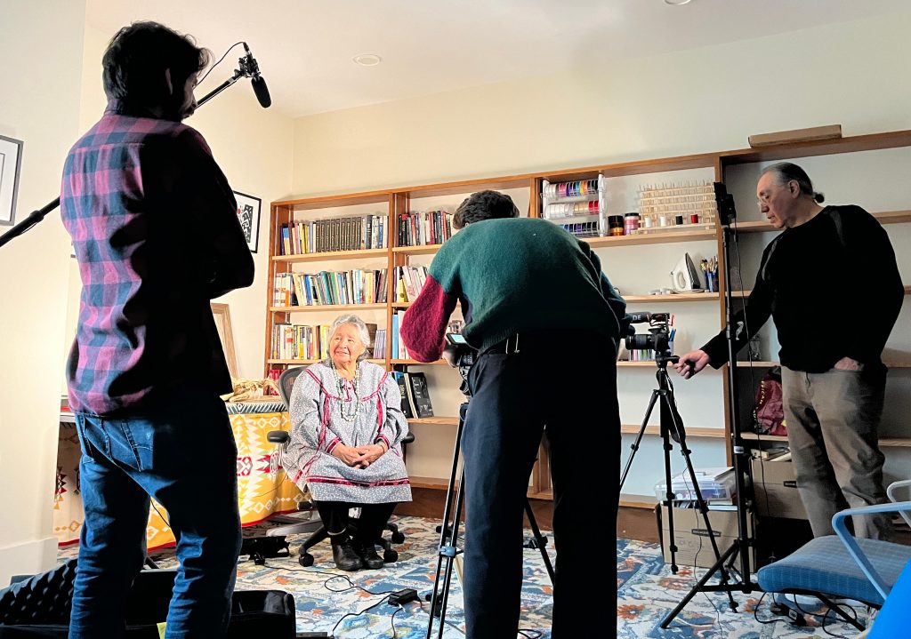 Person sitting in a chair in front a bookshelf with three camera operators facing her