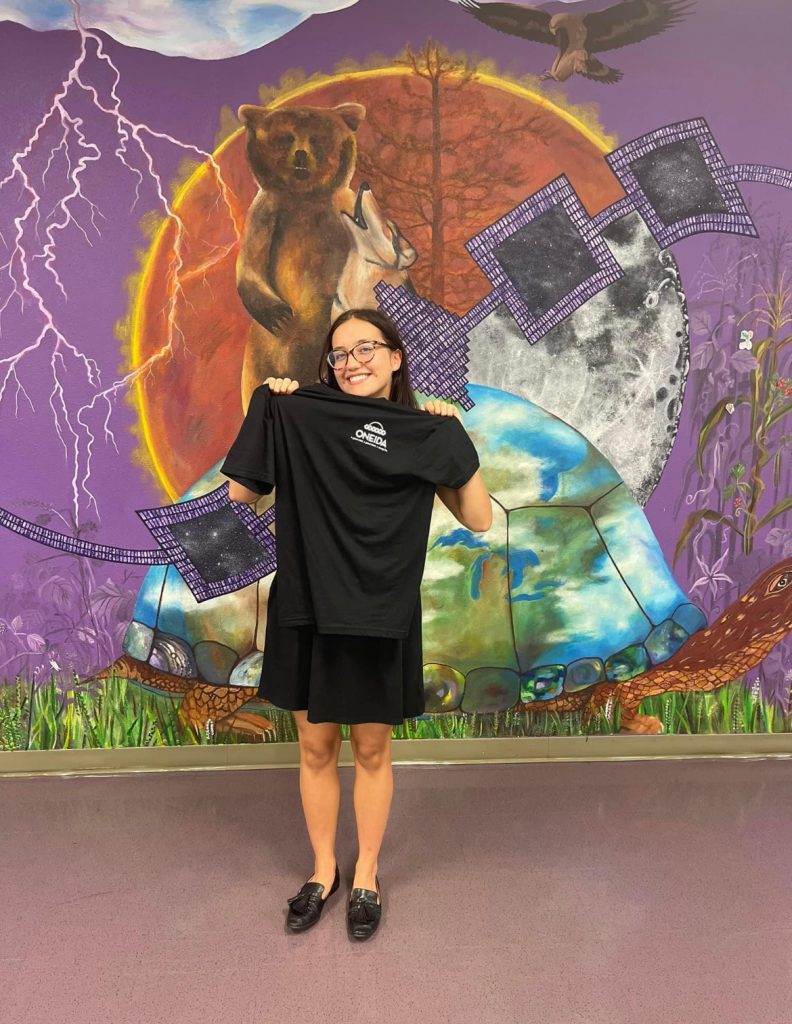Student standing in front of a wall with a mural holding up a black t-shirt