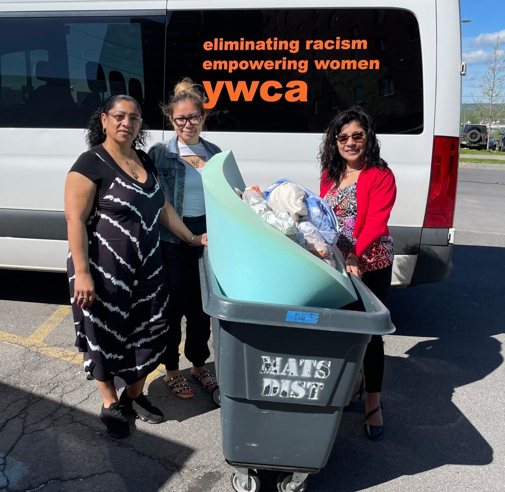Three people standing next to a white passenger van that says "eliminating racism, empowering women, YWCA" with a bin full of donated items
