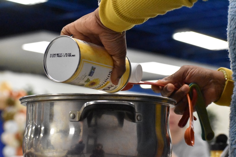 A person pouring sunflower oil into a measuring spoon over a silver pot.