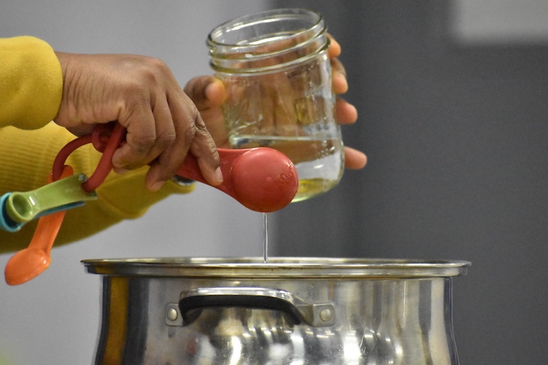 A person pouring a clear liquid from a measuring spoon into a silver pot. 