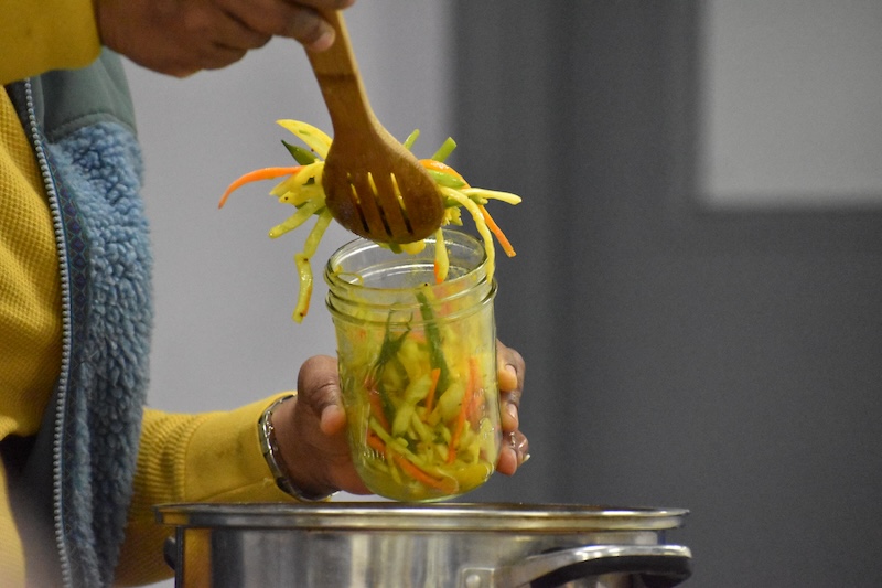 A person scooping a vegetable mixture with a wooden spoon into a glass jar.