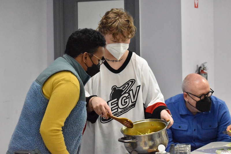 One person in a white hockey jersey, wearing a white mask stiring a mixture in a silver pot with a wooden spoon with an on looker in a black mask, looking at the mixture. 