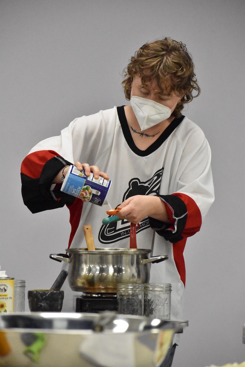 Person wearing a white hockey jersey and a white mask pouring salt into a measuring spoon over a silver pot.