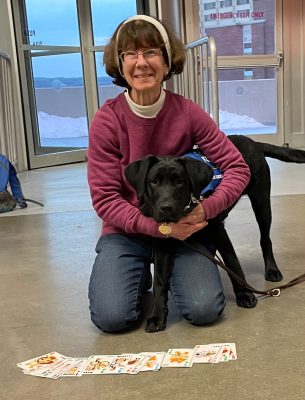 Women kneeling on the ground with their arms around a black dog with playing cards spread out in front of them