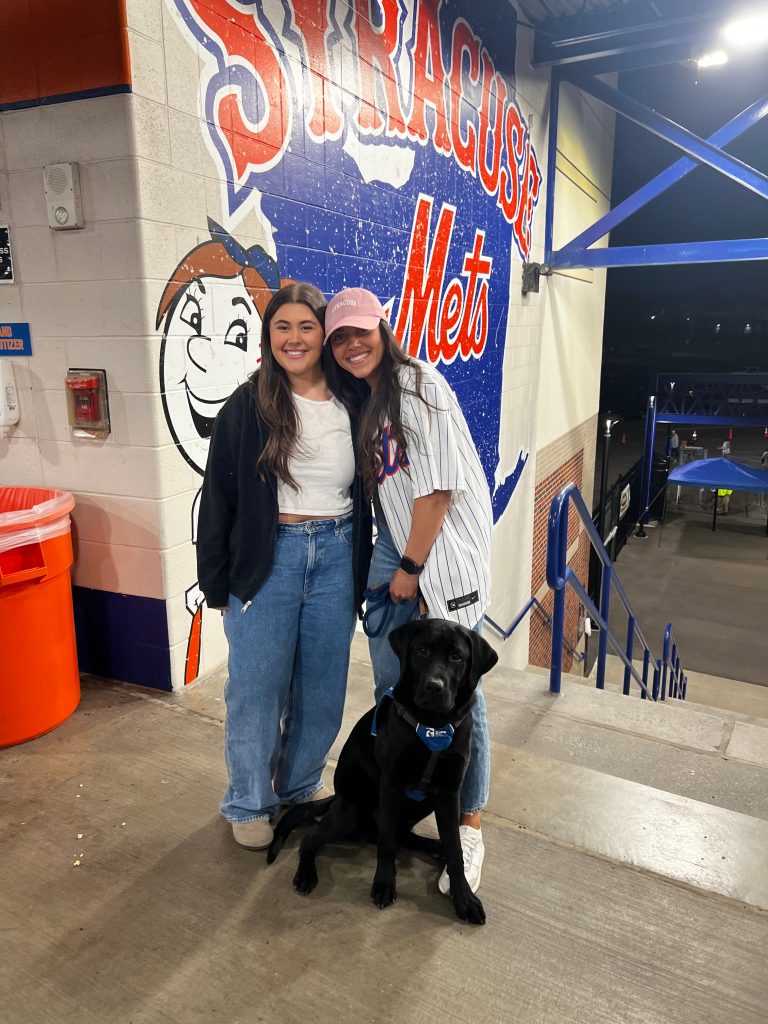 Two people standing together with a black dog standing right by them at the Syracuse Mets stadium.