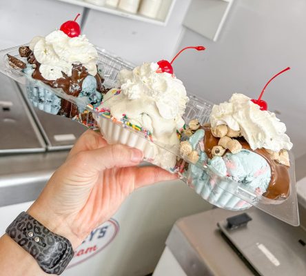 Hand holding a dish with three different scoops of ice cream with whipped cream and a cherry on top of each.