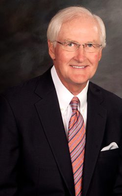 Portrait of a smiling person in a suit and striped tie against a dark background.
