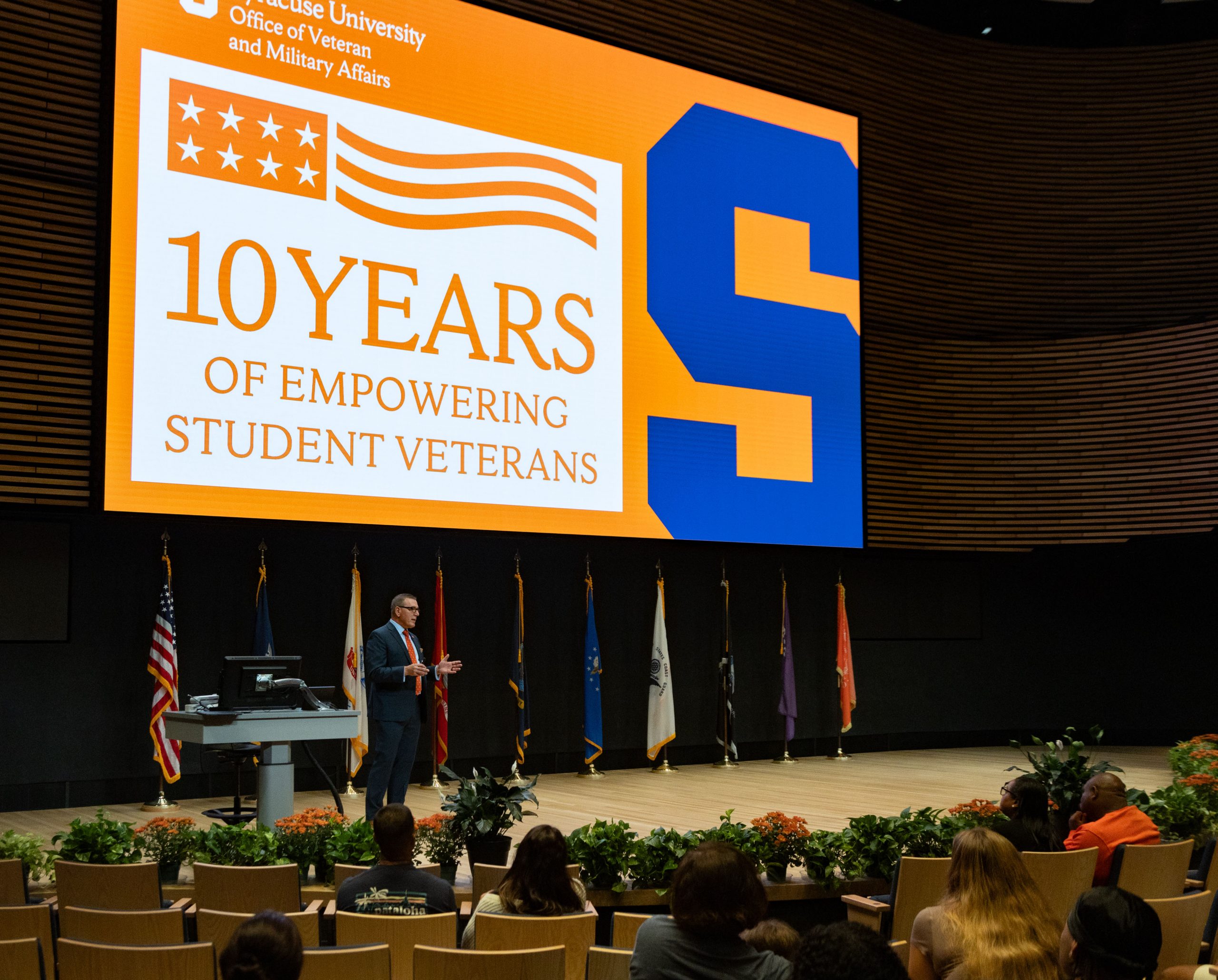 Person stands on a stage and speaks. In the background is a large screen with the inscription 