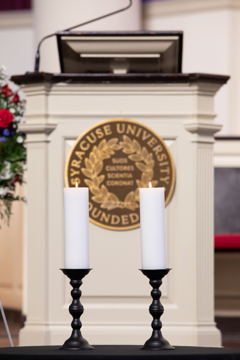 Two white candles lit in front of a podium with the Syracuse University seal on it.