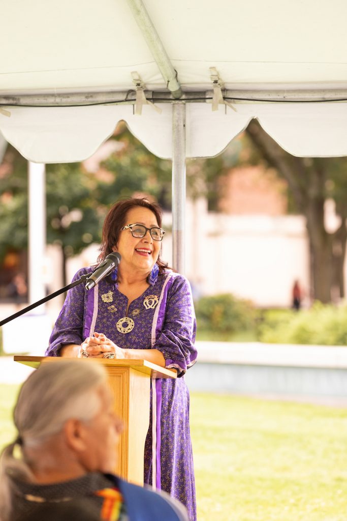 Woman in purple dress speaks at a podium under a tent