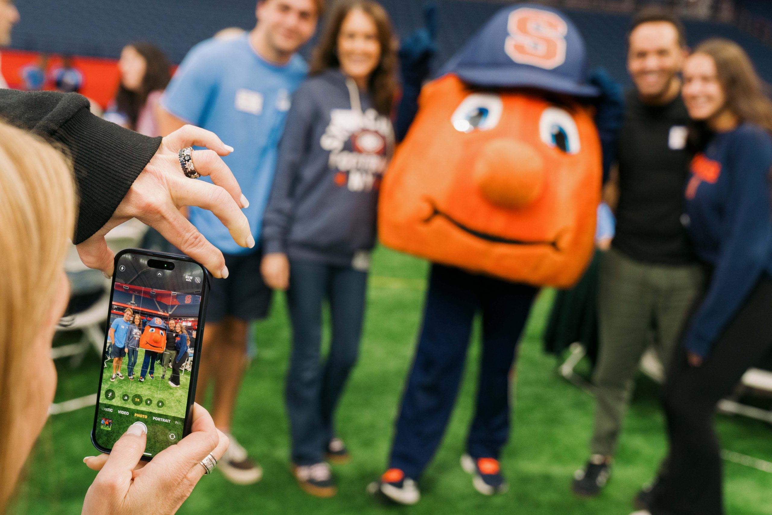 A group of people taking a photo with Otto while someone takes the photo with an iPhone. 