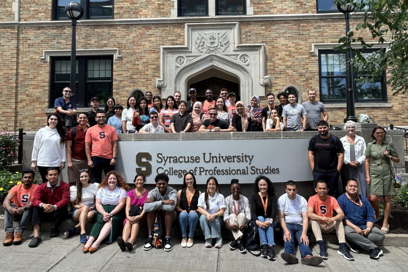 Large group of people gathered around the sign on the exterior of a building that says Syracuse University College of Professional Studies.