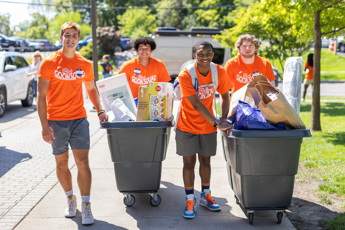 Four students in Good Squad t-shirts pushing grey bins full of items packed for move-in.