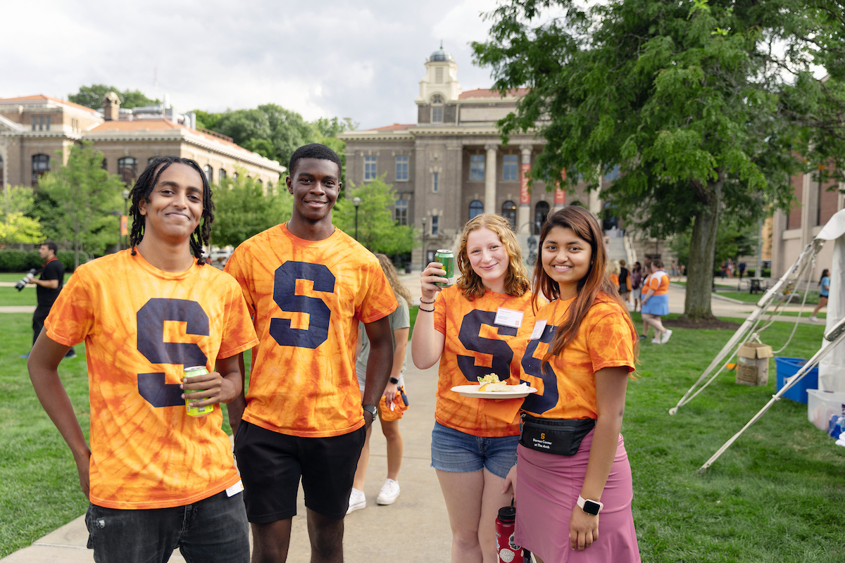 Four people standing together on the Quad all wearing orange t-shirts with a large blue S on them