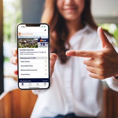 Person holding a smartphone displaying the Syracuse University OrangeNow app with weather, class schedule, and campus updates