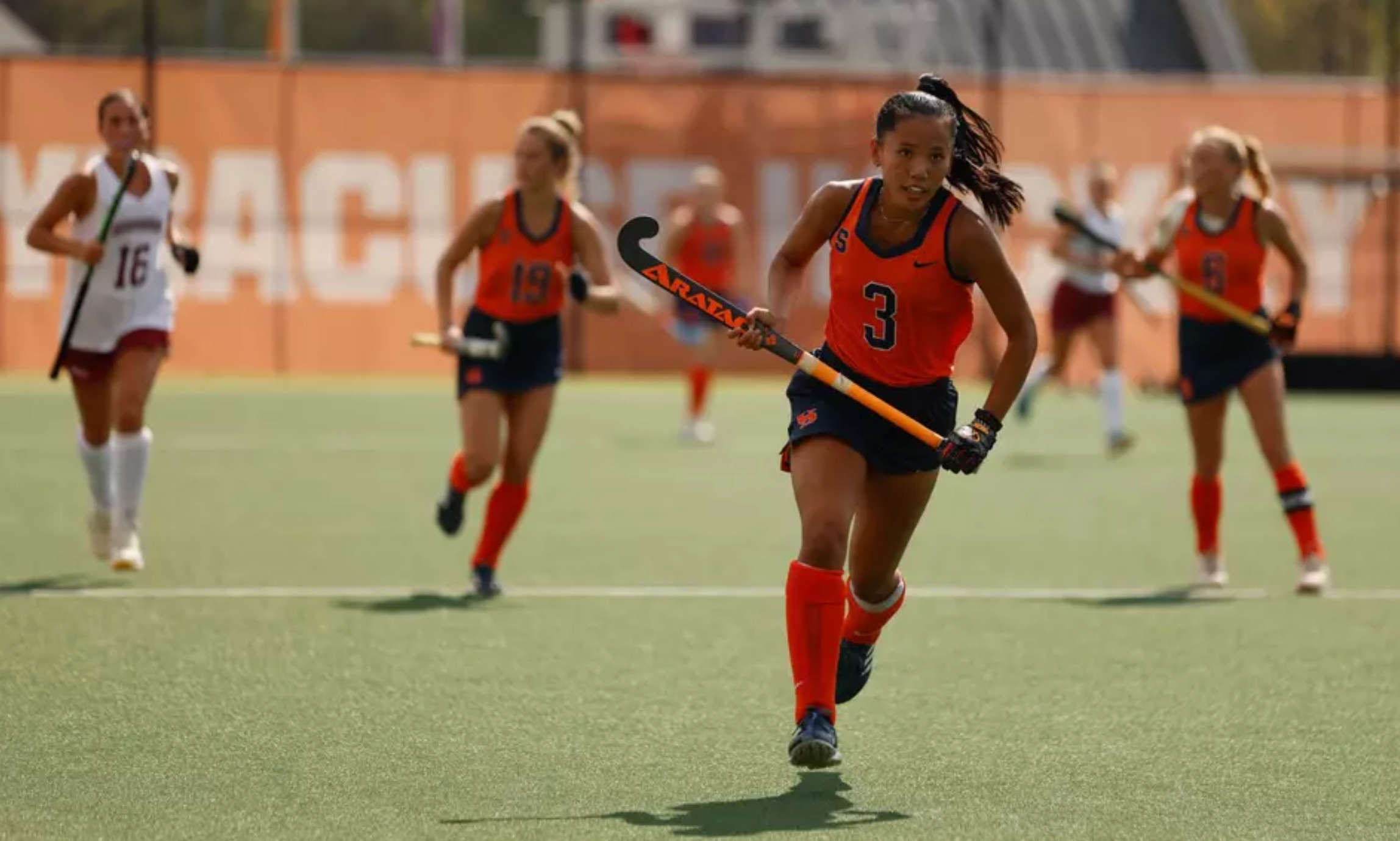 An athlete wearing number 3 and Syracuse University gear is playing field hockey, intensely focused while running with the ball. Other players are visible in the background on a sunny field.