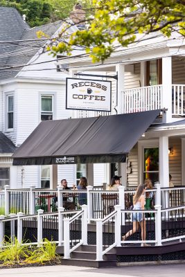 Customers enjoying drinks on the outdoor patio of Recess Coffee, a quaint café with a white porch and black awnings, nestled in a scenic neighborhood.