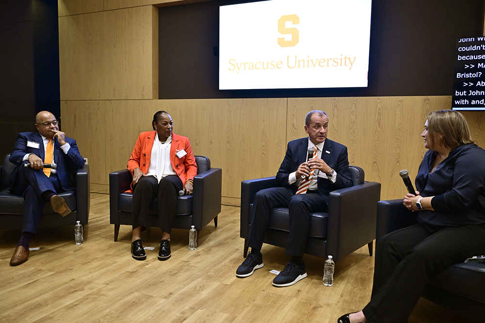 Four individuals are seated on a stage having a discussion at Syracuse University's new center in Washington, D.C.. A screen behind them displays the university's name and logo. One person holds a microphone and there are water bottles placed on the floor beside each chair.