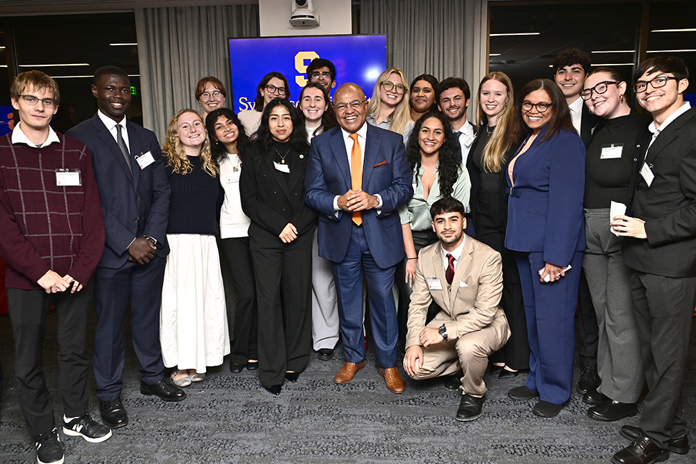 A group of smiling individuals dressed formally pose together in front of a blue digital screen with a Syracuse University logo.