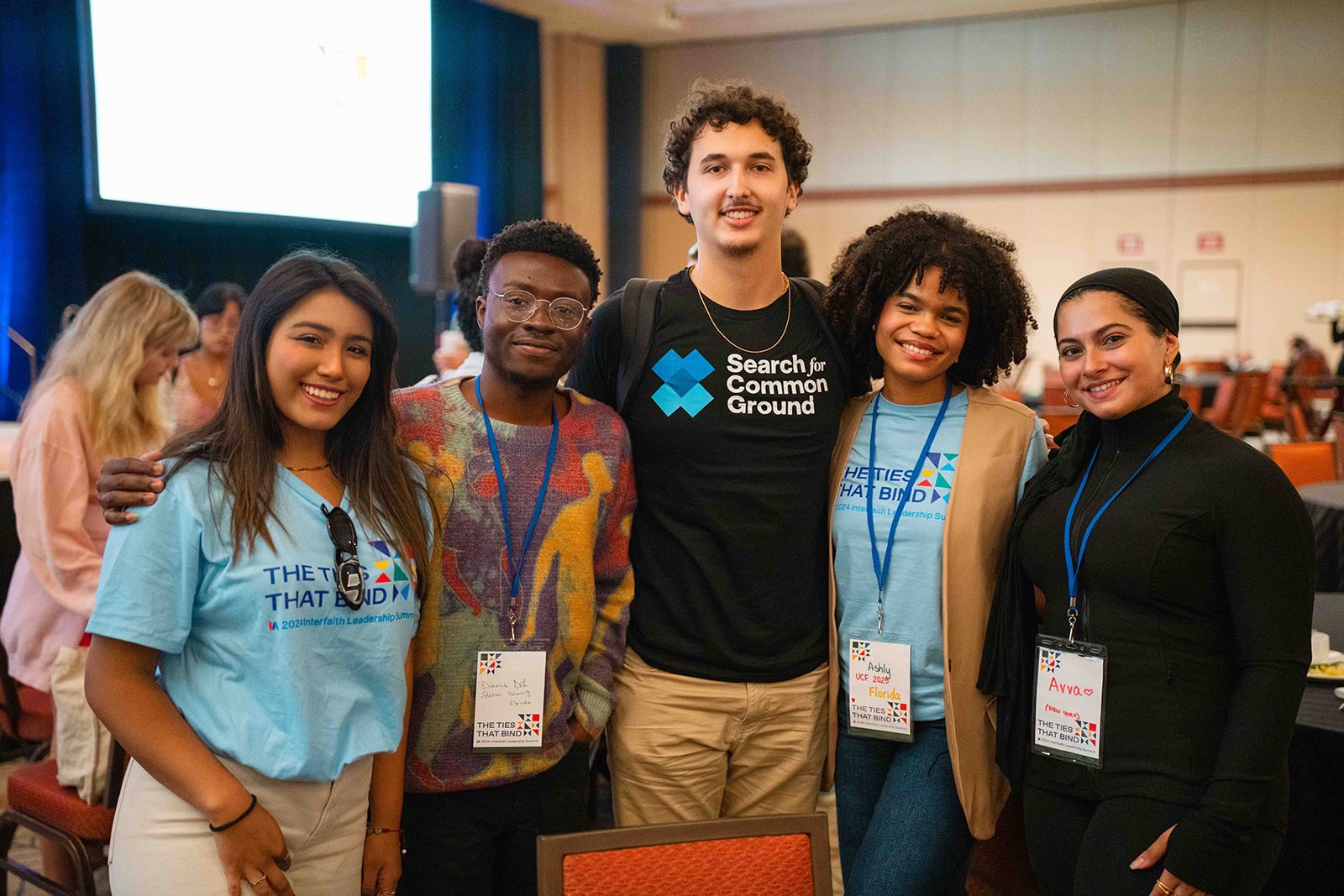 Five individuals wearing 'Search for Common Ground' T-shirts smiling together at a conference.