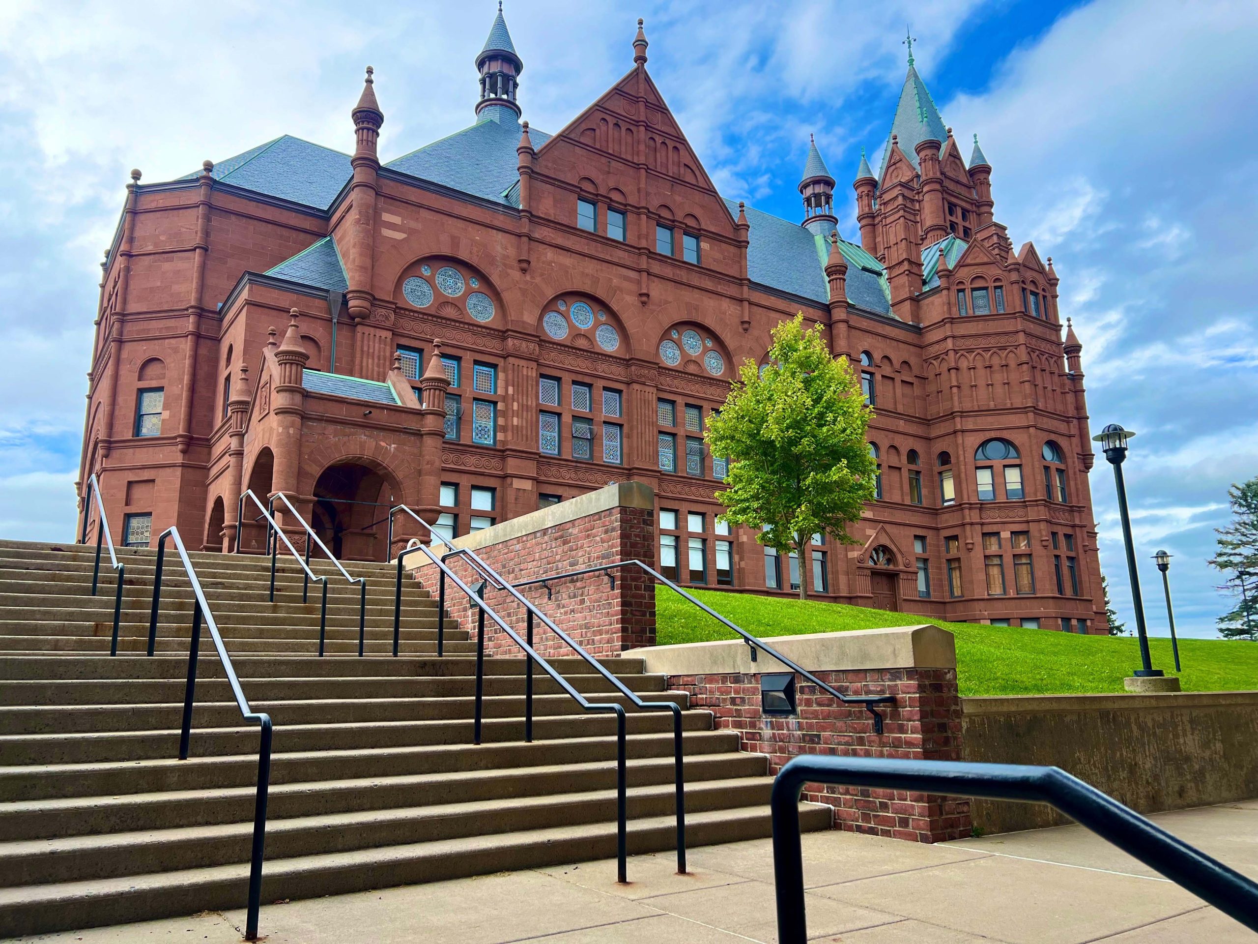 View of the Crouse College building at Syracuse University. The image features a large, ornate brick building with multiple turrets and arched windows, situated behind a wide staircase with metal railings, under a clear blue sky.