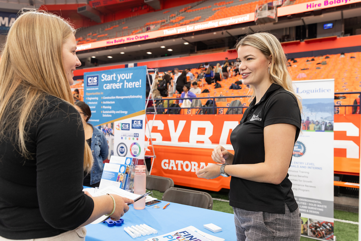 Two women engage in conversation at a career fair booth, one representing the company in a black polo shirt, while the other, holding documents, listens attentively. The booth features a sign that says, 'Start your career here!' and lists opportunities in engineering, architecture, and planning. The event takes place in a large, vibrant venue with orange bleachers and banners in the background.