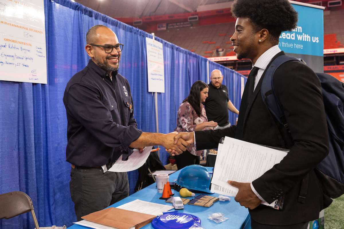 A career fair representative, dressed in a dark work shirt, smiles as he shakes hands with a young professional in a suit. The young man holds a resume and wears a backpack, while the booth displays a blue hard hat and various promotional items. In the background, additional attendees and recruiters are engaged in conversation, with blue curtains and company banners providing a backdrop for the event.