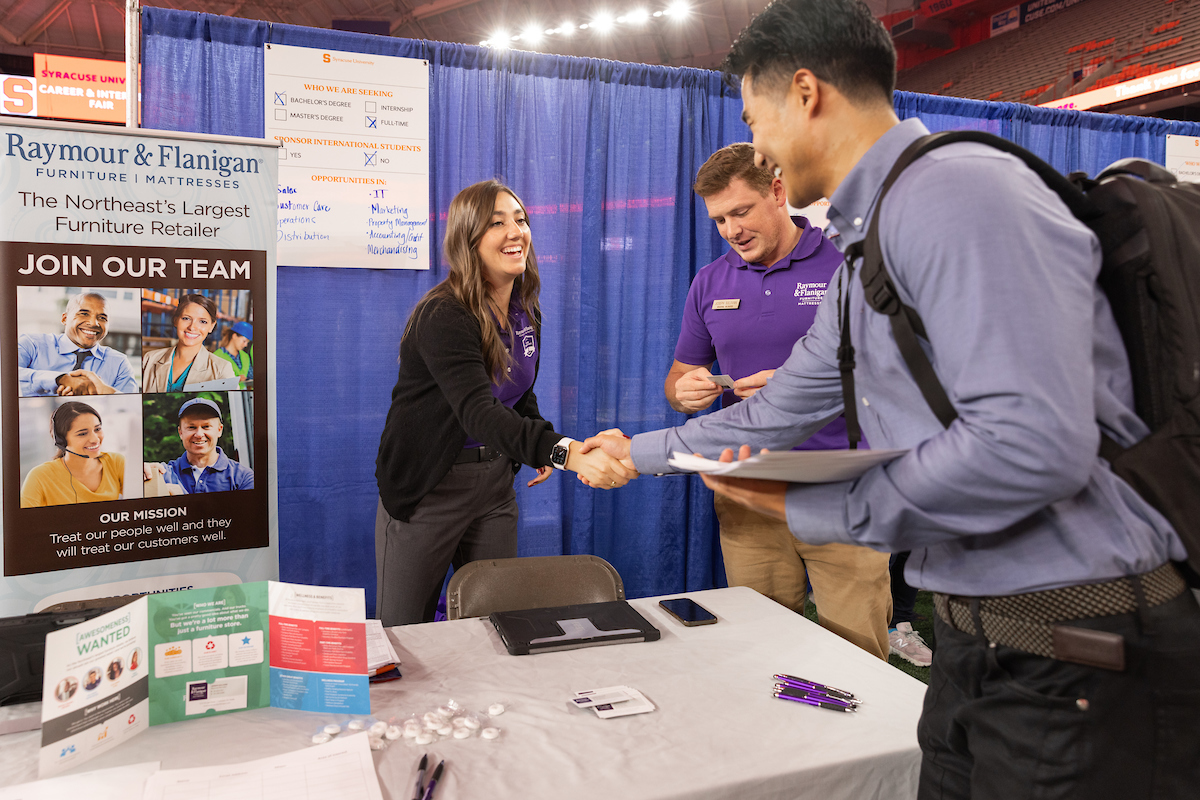 A young man shakes hands with a representative from Raymour & Flanigan at a career fair booth. The representative, along with her colleague wearing purple shirts, is smiling as they engage with the attendee. Behind them is a banner that reads 'Join our team' and highlights the company as the Northeast's largest furniture retailer. The booth displays various informational materials and brochures on the table, with a mission statement focused on treating their people and customers well.