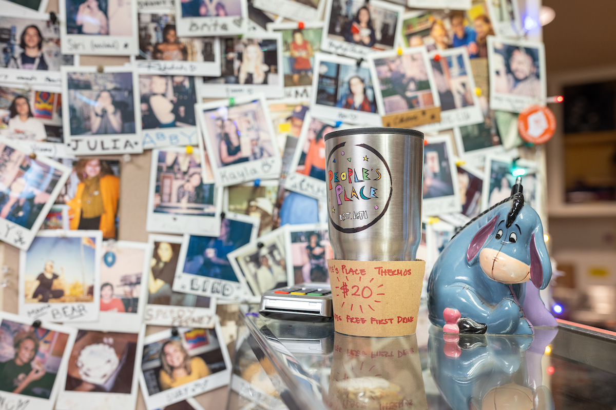 A coffee mug labeled "People's Place" surrounded by numerous Polaroid photos pinned to a wall, with a figurine of Eeyore from Winnie the Pooh sitting next to it.