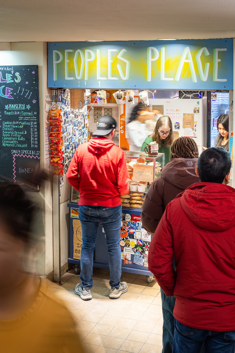 Customers browsing inside People's Place, a small store filled with various products including snacks and beverages. Some individuals are examining items, while others are at the checkout counter.