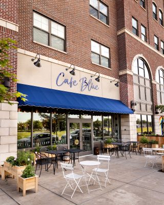 Exterior view of Café Blue, featuring a blue awning, white outdoor seating, and brick building with large windows.