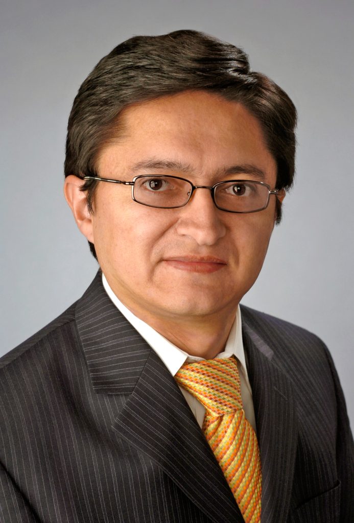 Portrait of an individual in a striped suit and a patterned tie, standing in front of a blurred background featuring a red-brick building and a statue.