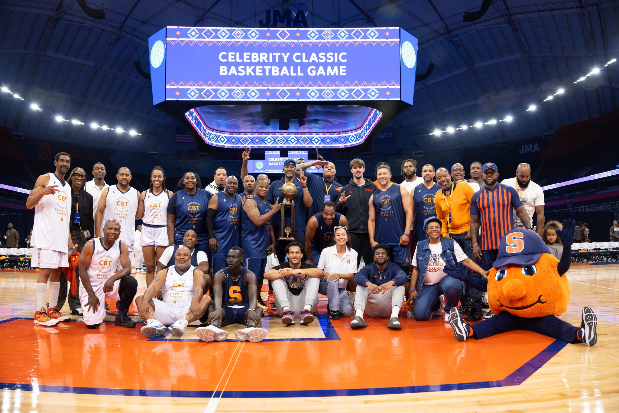 Group photo of participants at the Celebrity Classic Basketball Game, posing on a basketball court with a scoreboard reading "CELEBRITY CLASSIC BASKETBALL GAME" in the background, flanked by the Syracuse University mascot.