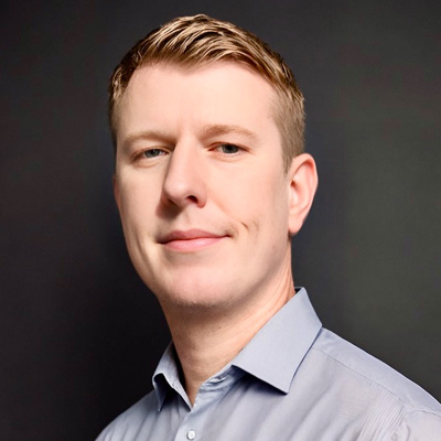 a man in a button-down shirt smiles in a studio headshot against a neutral backdrop