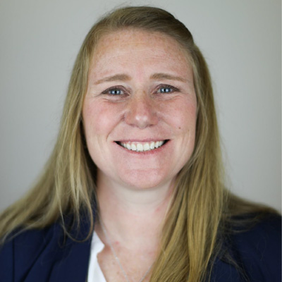 woman in white shirt and blue blazer smiles for a headshot against a neutral background