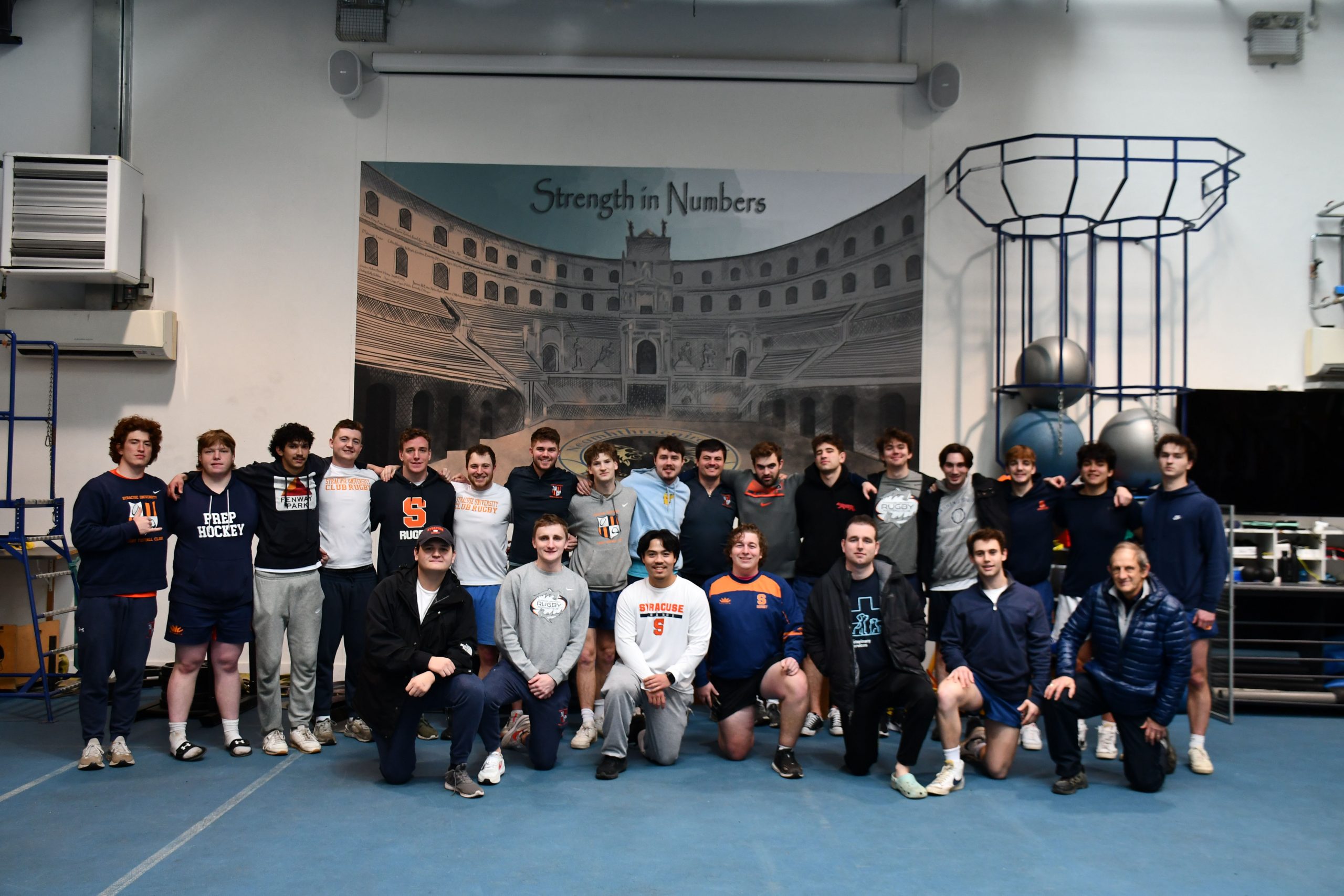 Group of athletes posing for a photo in a gym with a banner reading "Strength in Numbers" in the background.