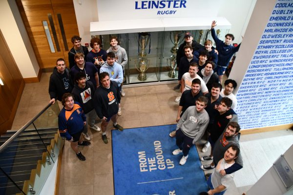 A group of people gathered in the Leinster Rugby headquarters, standing on and around a staircase, surrounded by trophies and rugby memorabilia.