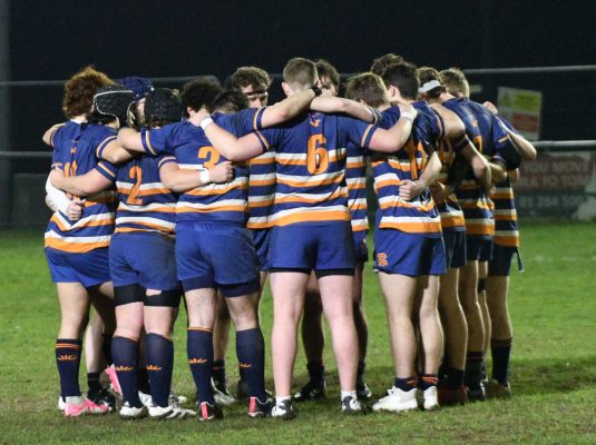 A group of Syracuse University rugby players in a huddle on the field at night, wearing orange and blue jerseys.