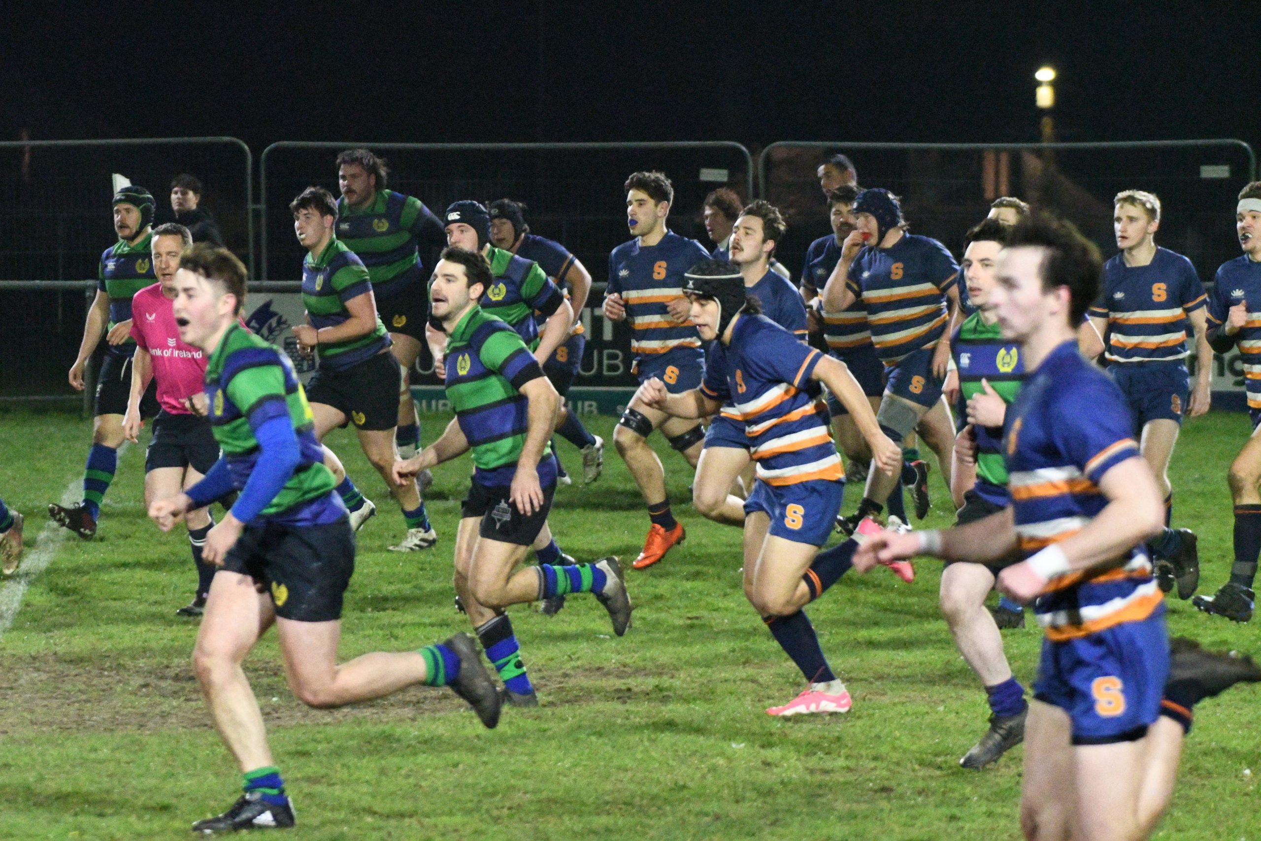 Players from two rugby teams, one in green and black stripes and another in blue and orange stripes, are actively competing in a night match on a well-lit field.