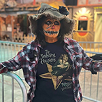 A person in skeleton makeup and a cowboy hat, wearing a "Rockin' Rodeos" t-shirt, standing with arms outstretched in an amusement park setting.