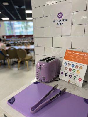 A gluten-free section in a cafeteria featuring a purple toaster on a purple mat, with tongs, and a sign labeled "Gluten Free Area." Next to it is a "Campus Dining Menu Labeling" guide with various food safety icons. In the background, numerous people are dining at tables.